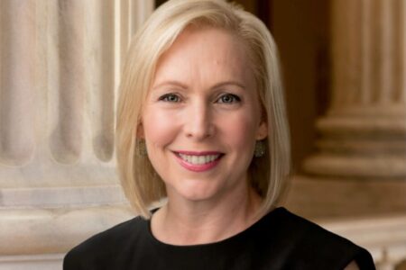 Senator Kirsten Gillibrand smiles, wearing a black top, with classical columns in the background.