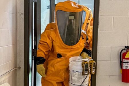 A firefighter in a full orange HazMat suit holds a bucket with detectors during a training drill.