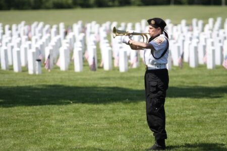 A bugler plays Taps at Sampson Cemetery during a Memorial Day ceremony, with rows of white headstones.