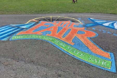Colorful chalk art of 'The Great New York State Fair' logo on asphalt, with grass and chairs in the background.