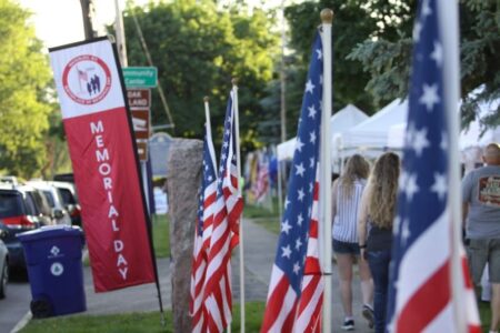 American flags and a Memorial Day banner line a street in Waterloo, NY, with people walking past tents at an event.