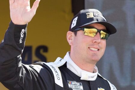 NASCAR driver Kyle Busch, wearing a racing suit and sunglasses, waves to a crowd.