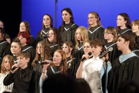Midlakes students perform in choir robes, singing into microphones on stage.