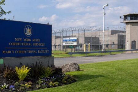 Entrance sign for Marcy Correctional Facility with a guard tower and barbed wire fence in the background.