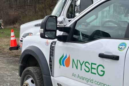 Side view of a white NYSEG utility truck with its logo, parked outdoors near another truck and a traffic cone.
