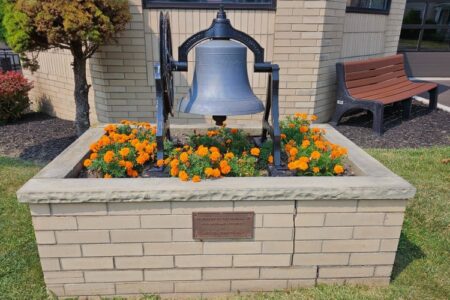 A large metal bell in a brick planter filled with bright orange flowers, with a bench and building in the background.