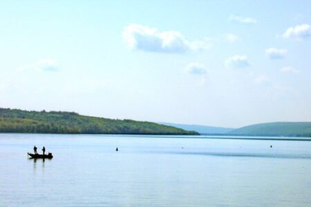 Two people fish from a boat on Owasco Lake with tree-covered hills under a blue sky.