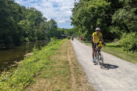 A person cycles on a sunny gravel path next to a canal, surrounded by green trees, with others in the distance.