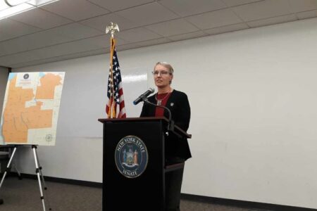 Senator Rachel May speaks at a podium with the NY State Senate seal, an American flag, and a map in the background.