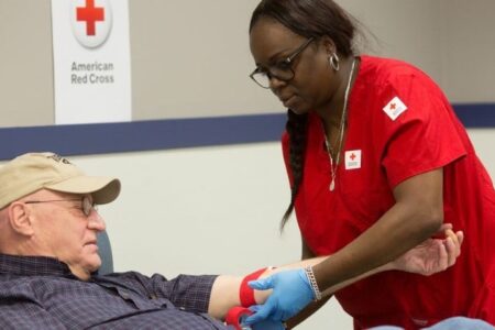 A Red Cross phlebotomist in red scrubs prepares an older man's arm for blood donation at a blood drive.