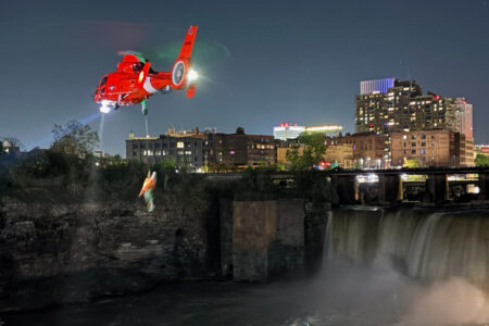 Coast Guard helicopter hoists person over Rochester's High Falls at night, city lights in background.
