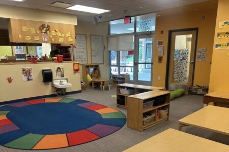An empty, colorful daycare classroom with a rainbow rug, small wooden tables, and a child-sized sink.