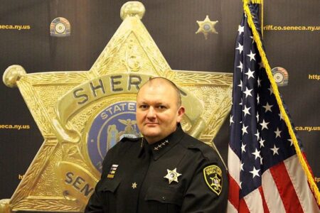Seneca County Sheriff Tim Thompson in uniform, standing before a large sheriff's star and American flag.