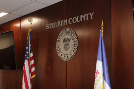 Steuben County seal and name on a wooden wall, flanked by the American flag and county flag.