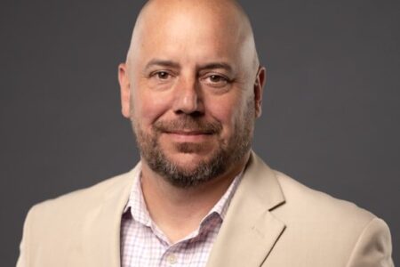 Headshot of Steve Griffin, a bald man with a beard, wearing a tan jacket, smiling against a gray background.