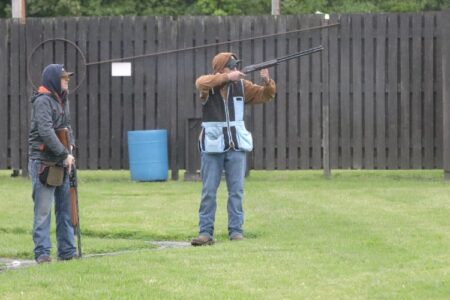 Two men at a trap shooting range, one aiming a shotgun, the other standing by with a shotgun.
