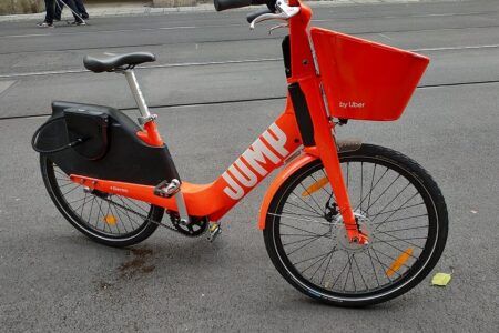 An orange JUMP by Uber electric bicycle with a front basket is parked on a paved surface.
