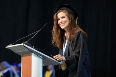 A woman in a graduation cap and gown smiles while speaking at a podium with microphones.