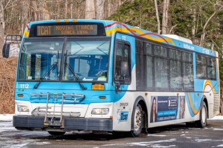 A blue and white TCAT bus is parked on a snowy road with bare trees in the background.