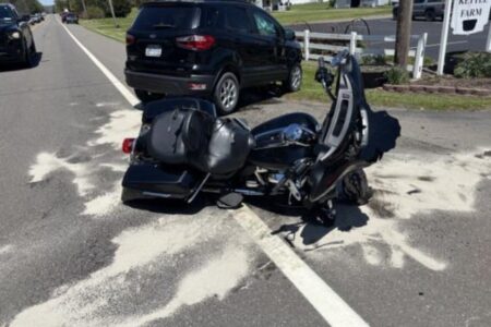 A black motorcycle lies on its side on a road next to a black SUV, with absorbent material on the pavement.