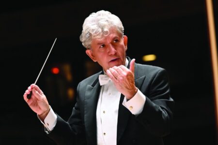 A male conductor with gray hair in a tuxedo holds a baton and gestures during a performance.