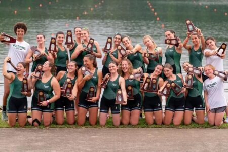 Herons Rowing team celebrates 3rd place at NCAAs, posing with numerous trophies by the water.