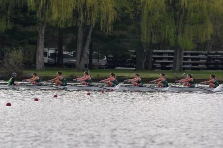 A women's rowing team in a long boat with a coxswain, racing on a lake with trees in the background.