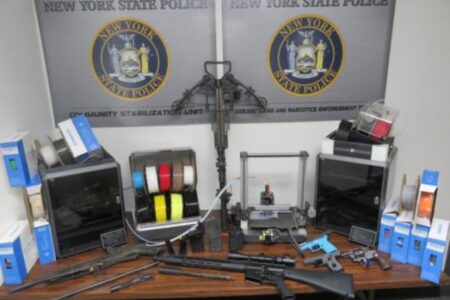 Seized firearms, 3D printers, and filament on a table with New York State Police signs in the background.
