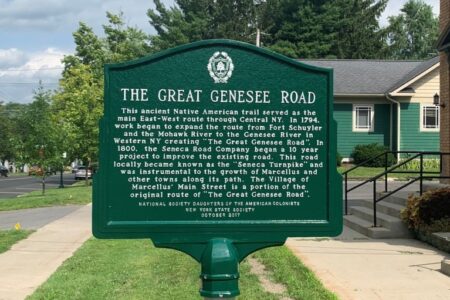 A green historical marker for 'The Great Genesee Road' stands on a sidewalk, detailing its history as a Native American trail.