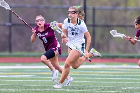 William Smith lacrosse player Sypeck runs with the ball on the field, pursued by a Union opponent.