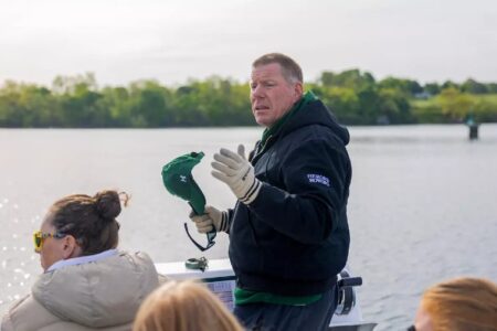 Rowing coach speaks on a boat, holding a green hat, with water and trees in the background.