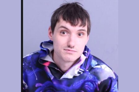 Mugshot of a young man with brown hair and a light mustache, wearing a blue and purple galaxy-print hoodie.