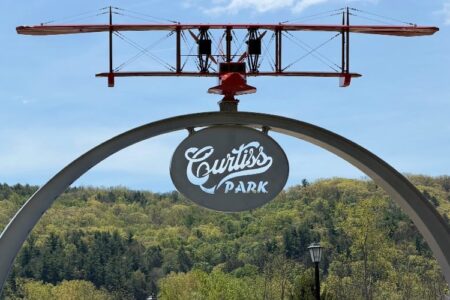 Sign for Curtiss Park with a red biplane model above it, against a backdrop of green hills and blue sky.