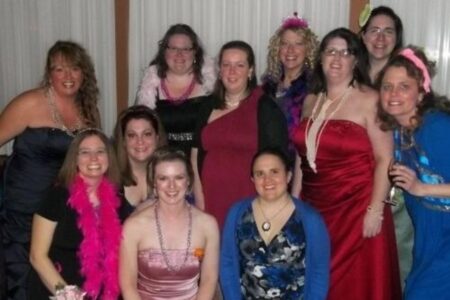 A group of smiling women in formal dresses and accessories pose together at the CNY Mom Prom.
