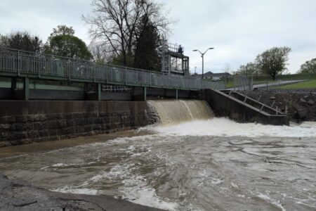 Water flows over a dam on the Owasco River under an overcast sky, with a bridge above.