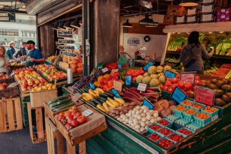 A vibrant farmers market stall overflows with colorful fresh fruits and vegetables, with shoppers in the background.