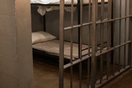 A stark prison cell featuring metal bunk beds with white bedding and vertical metal bars in the foreground.