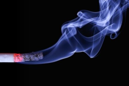 A lit cigarette with glowing embers and blue smoke against a black background.