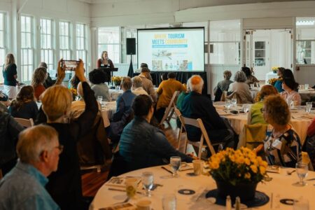 Attendees at the Tour Cayuga 2nd Annual Community Luncheon, with a speaker at a podium and a screen showing 'Where Tourism Meets Community'.