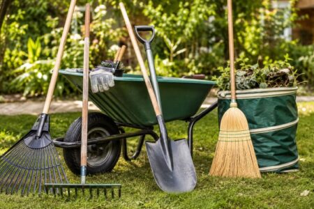 Gardening tools including rakes, shovel, wheelbarrow, and broom on a lawn with a full yard waste bag.