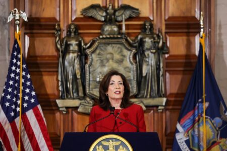Governor Kathy Hochul speaks at a podium with the New York State Seal and flags behind her.