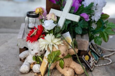 A somber memorial featuring teddy bears, flowers, a white cross, and a candle, honoring victims of the Uvalde tragedy.