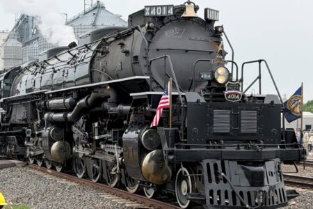Union Pacific Big Boy No. 4014 steam locomotive with smoke, flags, and a crowd watching near grain silos.