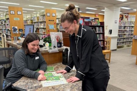 Two library staff members smiling at a counter, looking at papers or a game board.
