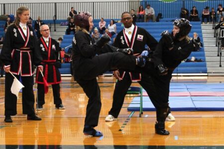 Two martial artists in black uniforms spar during a competition, one delivering a high kick.