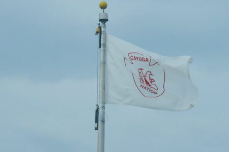 The white flag of the Cayuga Nation with its red emblem and text flies on a flagpole against a light blue sky.