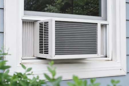 A window air conditioner installed in a light blue house window with green foliage in the foreground.