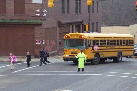 A crossing guard in a bright green coat helps students cross the street in front of a yellow school bus.