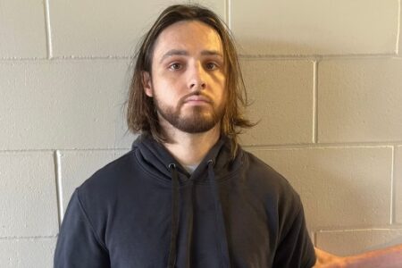 A man with long brown hair and a beard, wearing a dark hoodie, stands against a light-colored cinder block wall.