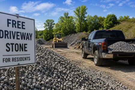 A sign reads 'FREE DRIVEWAY STONE FOR RESIDENTS' next to piles of gravel, a bulldozer, and a truck loaded with stone.